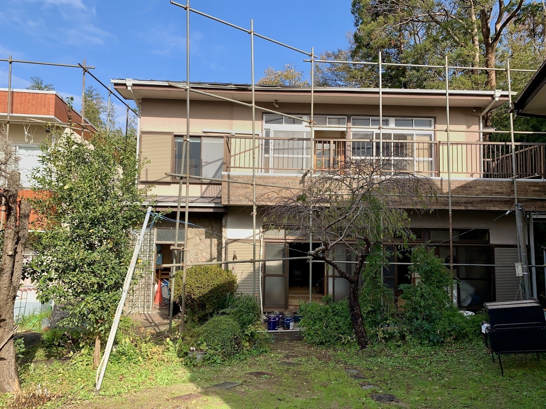 An old tan and brown Japanese style house with scaffolding around prior to it being torn down. Yard is kind of overgrown. There’s a makeshift utility pole with an electric wire strung from it. 