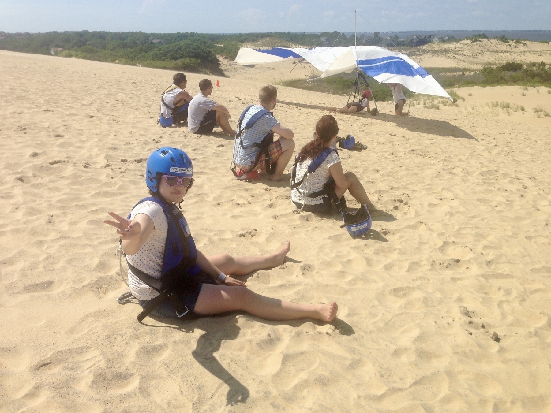 Young woman giving a peace sign sitting on the sand dunes at Kitty Hawk NC, before her hang glider flight. 